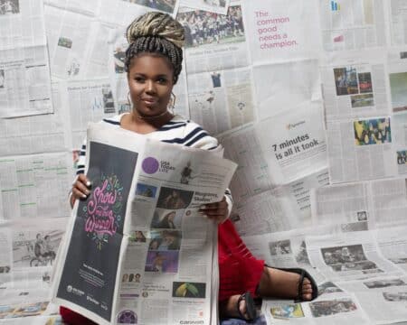 Femme lisant un grand journal entourée de feuilles de presse, actualité sociale dans un magazine.