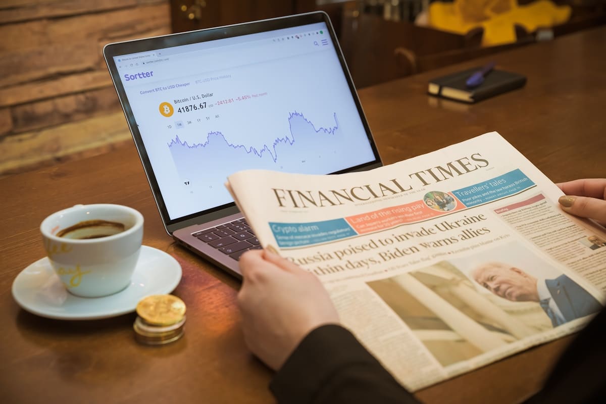 Ordinateur portable affichant un graphique de crypto-monnaie, journal financier, tasse de café et pièces de monnaie.