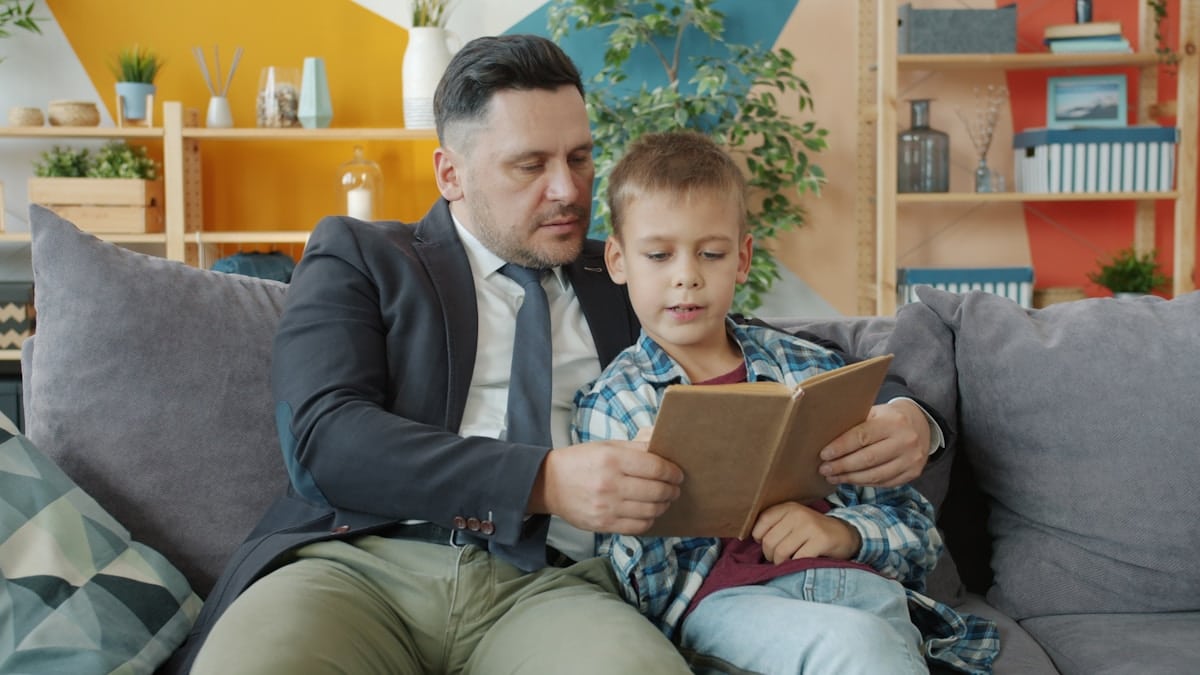 Un homme en costume lisant un livre avec un jeune garçon sur un canapé.