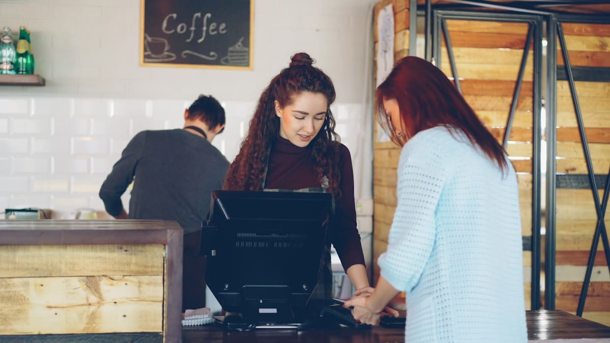 Deux jeunes femmes travaillant ensemble dans un café, avec un homme en arrière-plan.