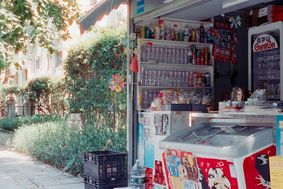 Épicerie de quartier avec étagères remplies de produits, située dans un environnement verdoyant.