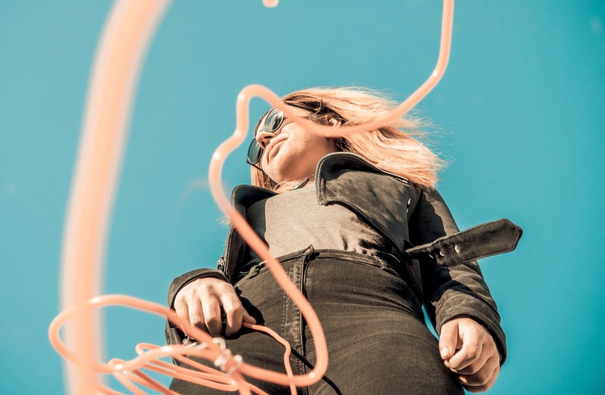 Femme blonde aux cheveux ébouriffés, portant des lunettes de soleil, assise sur une chaise.