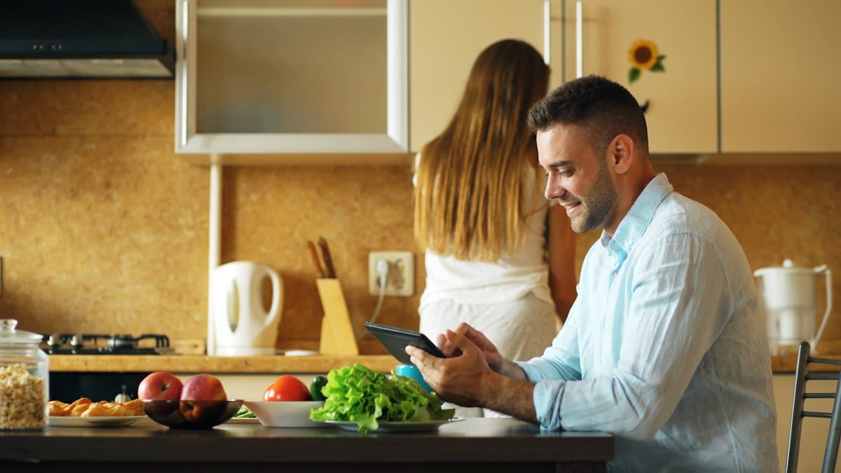 Couple dans une cuisine, homme utilisant une tablette numérique.